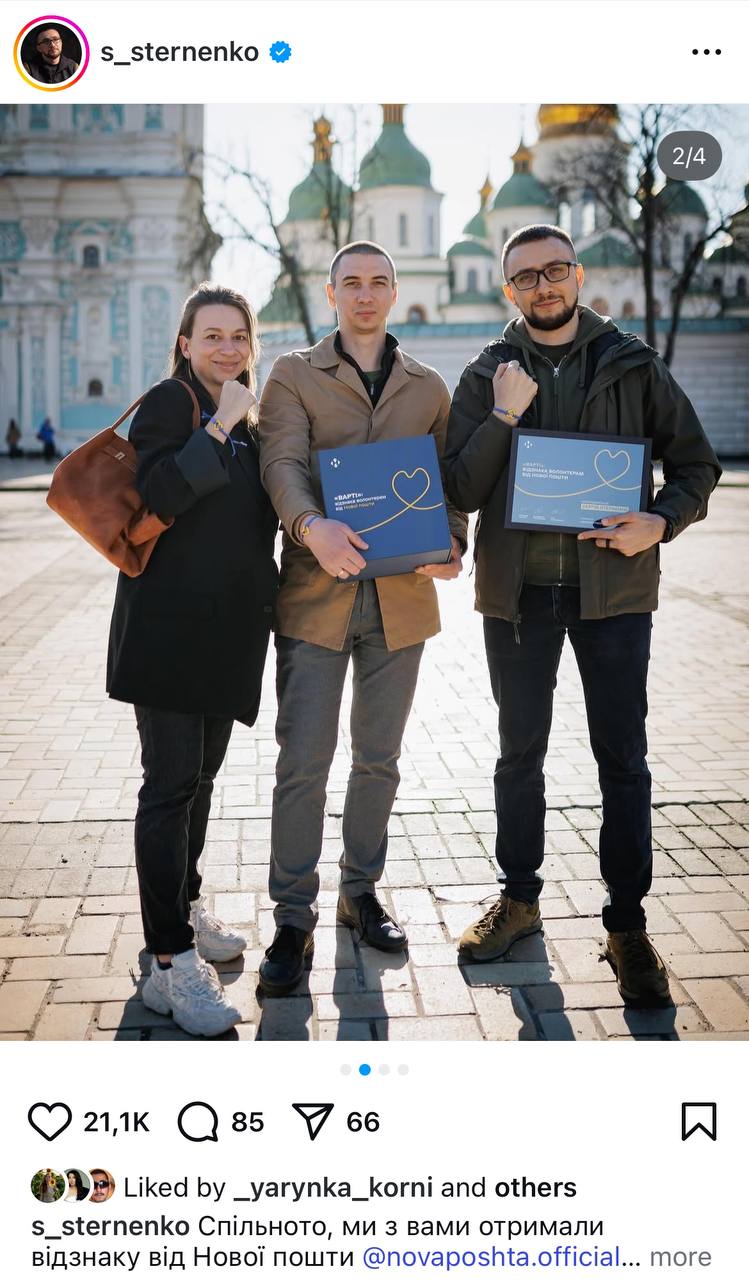Три волонтери позують на вулиці, тримаючи нагороди з відзнакою "Варті" від Нової пошти на фоні архітектурної будівлі.