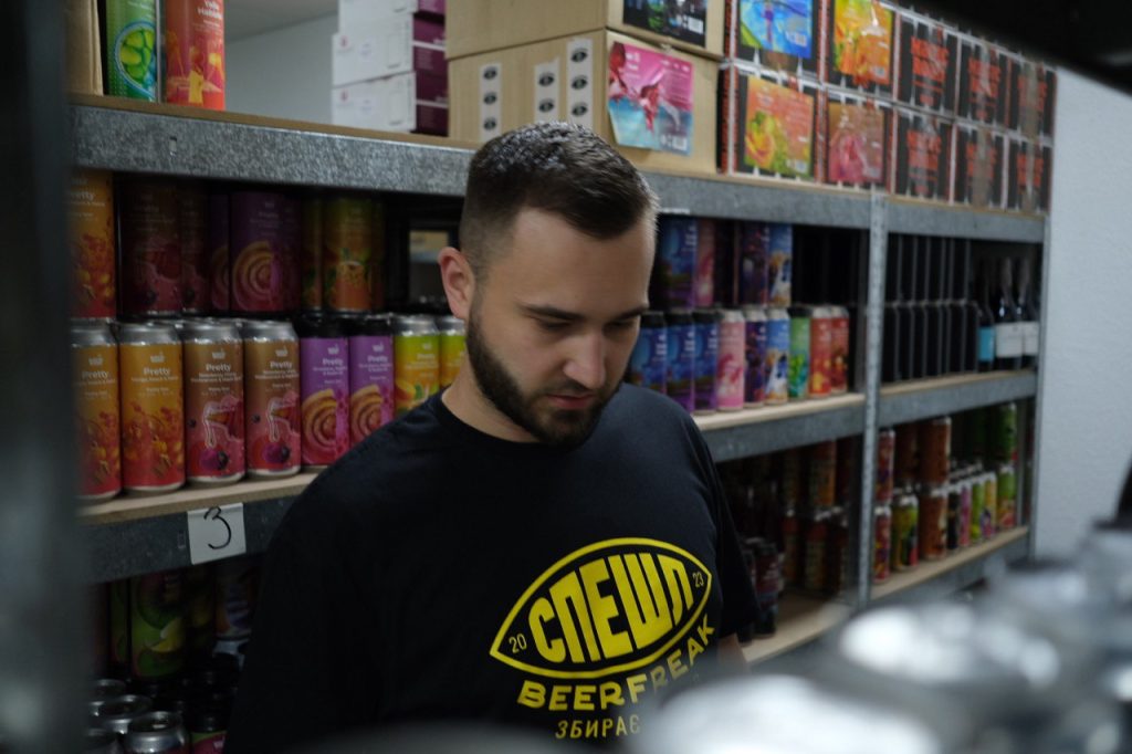 Чоловік у чорній футболці з логотипом BeerFreak, який стоїть на фоні полиць з банками та пляшками пива.