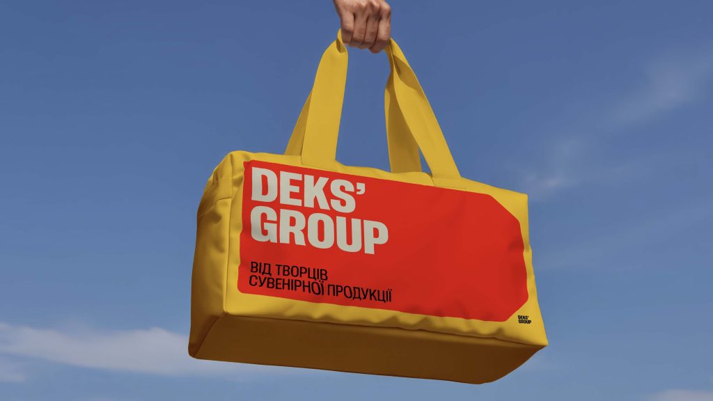 Яскрава жовта сумка з червоною наклейкою на якій написано 'DEKS' GROUP' та українським текстом 'ВІД ТВОРЦІВ СУВЕНІРНОЇ ПРОДУКЦІЇ', на фоні голубого неба.