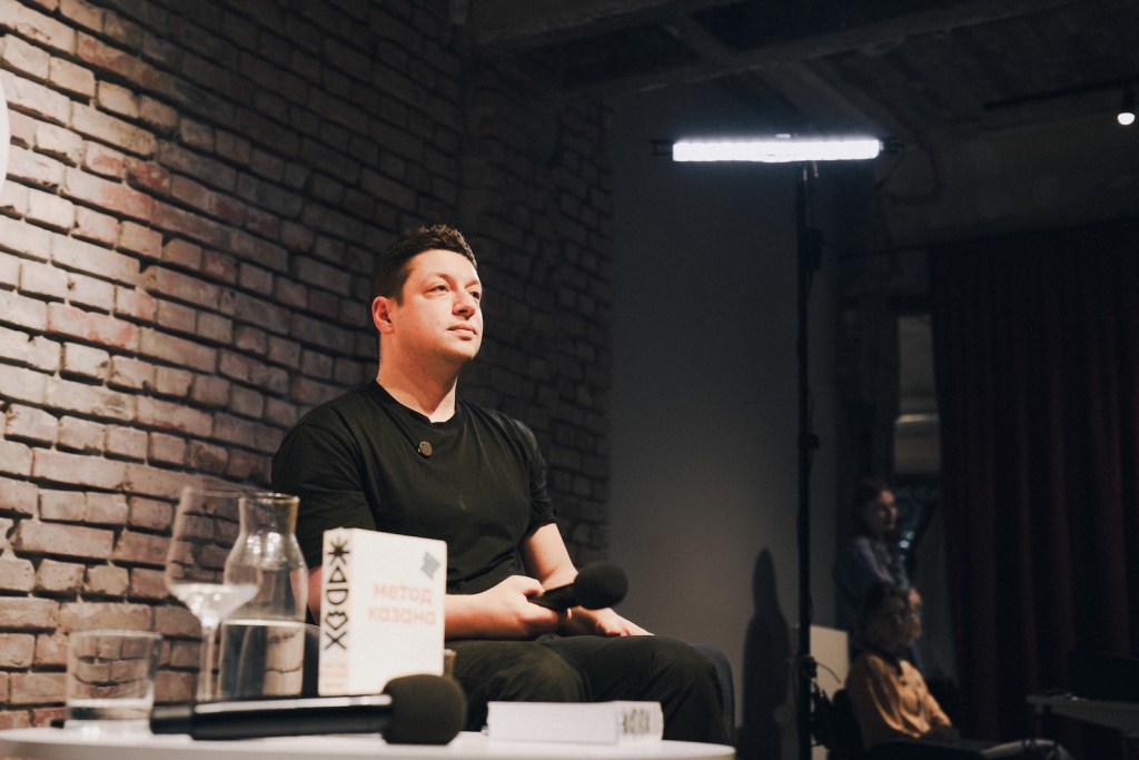 Чоловік сидить на сцені під час заходу. На фоні бачимо цегляну стіну. На столі перед ним є склянка та книга з назвою 'Метод Казано'.